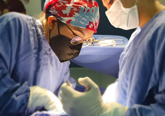 Dra. Irasema Salgado Falcón con bata azul, gorro, lentes y cubrebocas negro, operando a paciente