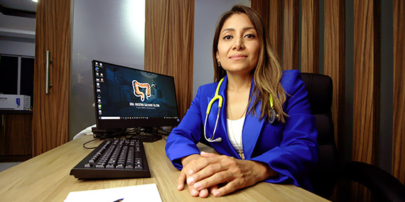 Foto de la Dra. Irasema Salgado Falcón con traje saco azul y estetoscopio, sentada en su escritorio junto a su computadora